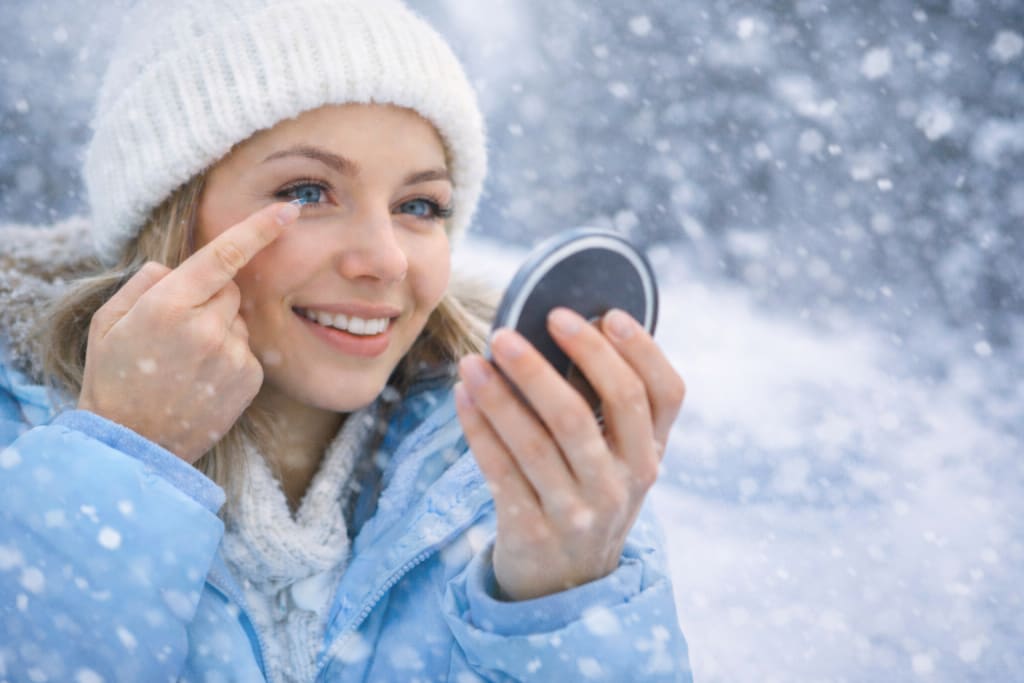 Woman wearing contact lenses outdoors in snowy winter weather, applying a lens while holding a compact mirror.