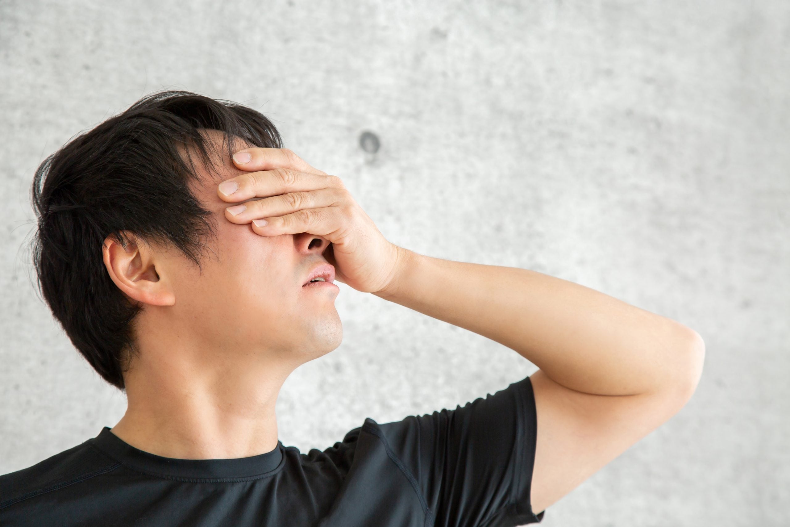 Man in a black shirt covers his eyes with his hand, showing discomfort or eye strain against a gray wall.