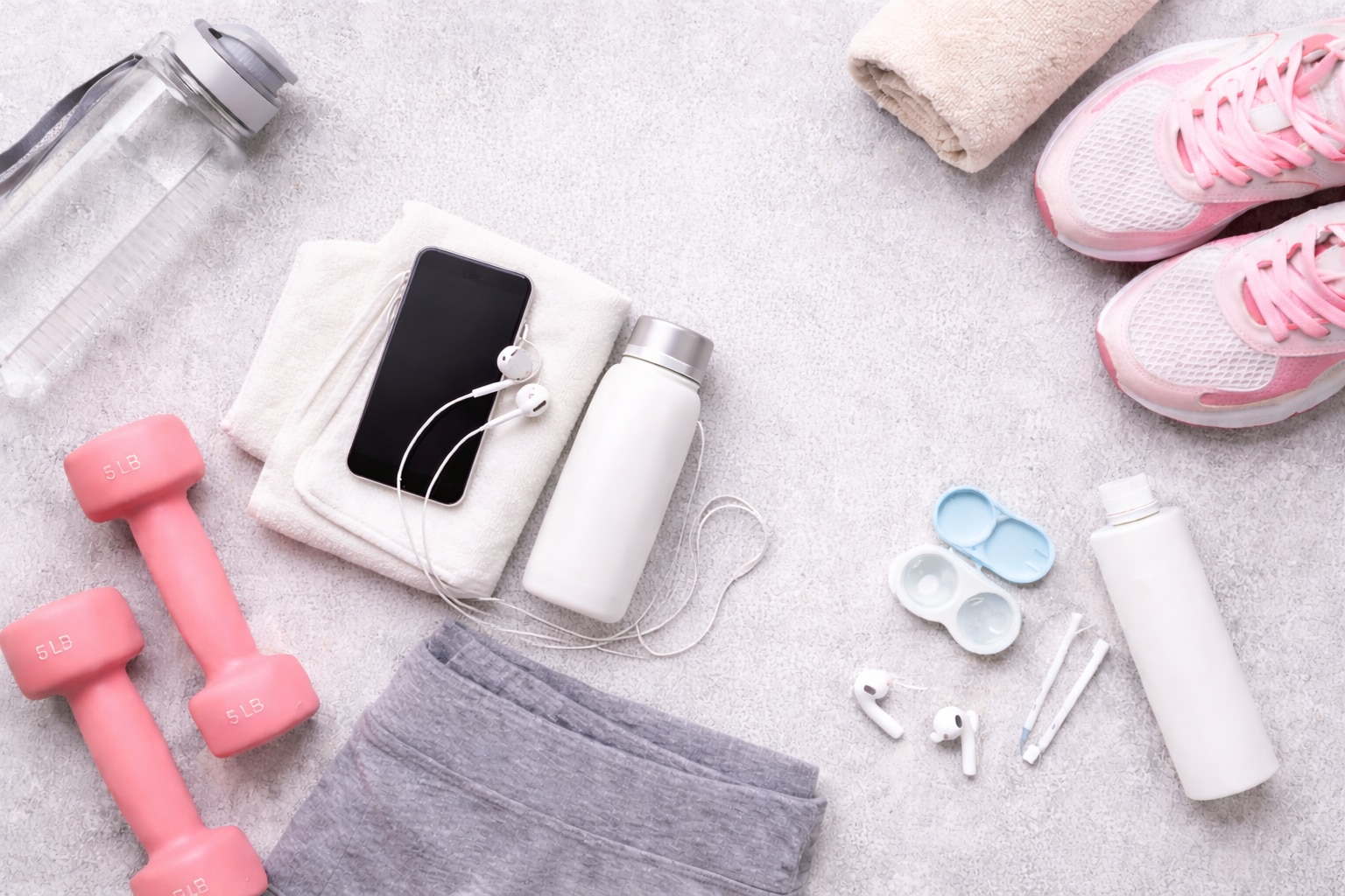 Flat lay of gym and contact lens essentials on a light gray background, including pink dumbbells, a clear water bottle, pink sneakers, a beige towel, gray workout shorts, a smartphone with earbuds, an open contact lens case, and a contact lens solution bottle.