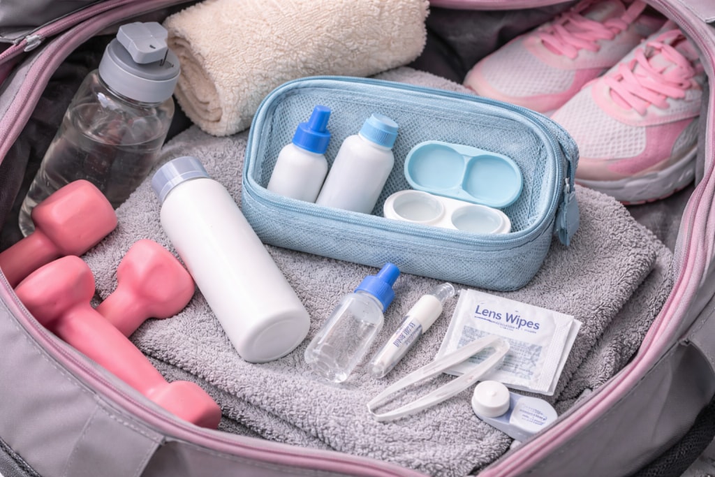 Open gym bag packed with a contact lens care kit, including rewetting drops, lens solution, a contact lens case, lens wipes, tweezers, and a towel, alongside pink dumbbells, a water bottle, and sneakers.