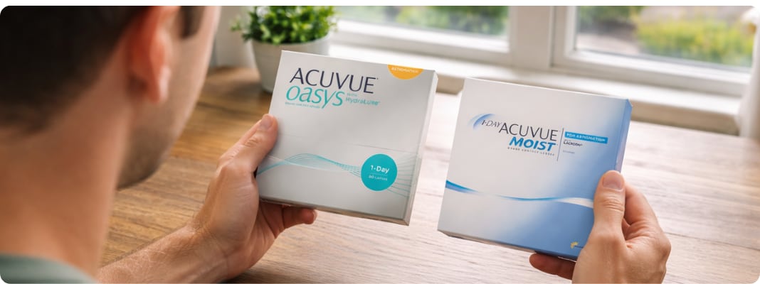 Man viewed from behind comparing Acuvue Oasys 1-Day vs Acuvue Moist for Astigmatism boxes on a wooden table near a window with a blurred garden outside.