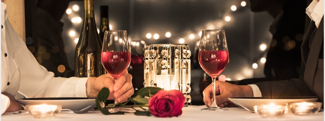 Couple seated at a candlelit dinner table holding glasses of red wine, with a rose and string lights in the background.