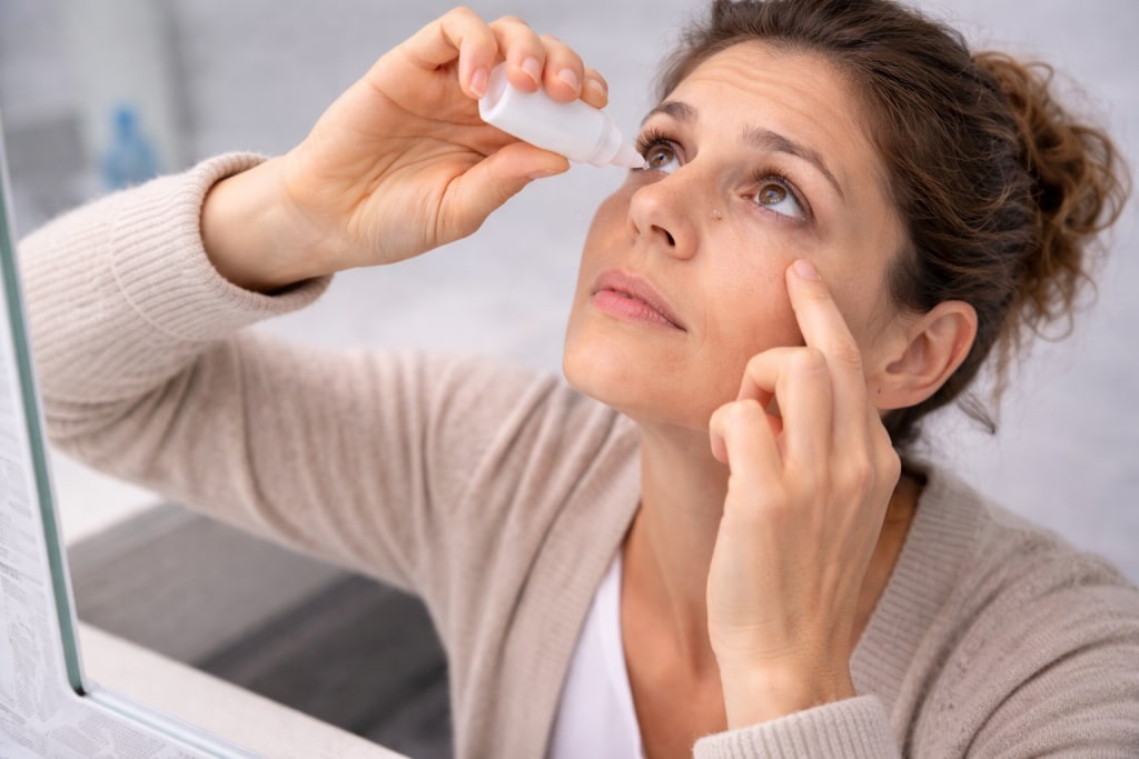 Woman applying rewetting drops while looking in a mirror, illustrating Contacts Blurry Causes and Fixes for dryness-related blurry vision with contact lenses.